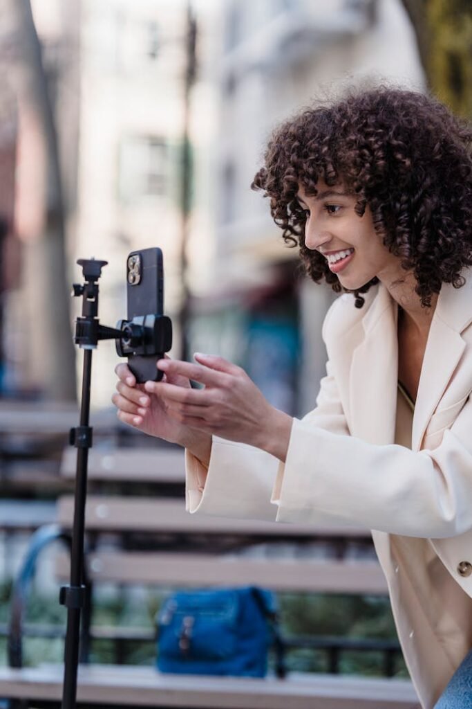 243203 Smiling woman with curly hair uses smartphone and tripod for vlogging outdoors.